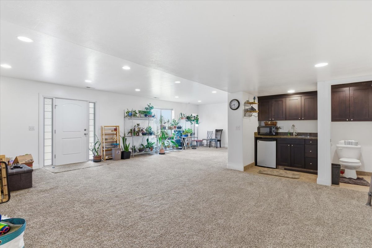 Indoor wet bar with light colored carpet, dark wood finish cabinets, refrigerator, recessed lighting, and dark countertops