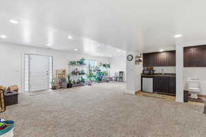Indoor wet bar with light colored carpet, dark wood finish cabinets, refrigerator, recessed lighting, and dark countertops