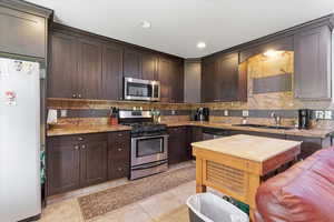 Kitchen featuring stainless steel appliances, dark wood finish cabinets, light stone countertops, light tile patterned floors, and tasteful backsplash