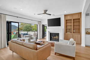 Living room with light wood finished floors, a ceiling fan, a glass covered fireplace, built in features, and a textured ceiling