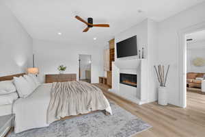 Bedroom featuring ensuite bath, light wood-style flooring, a ceiling fan, and a glass covered fireplace