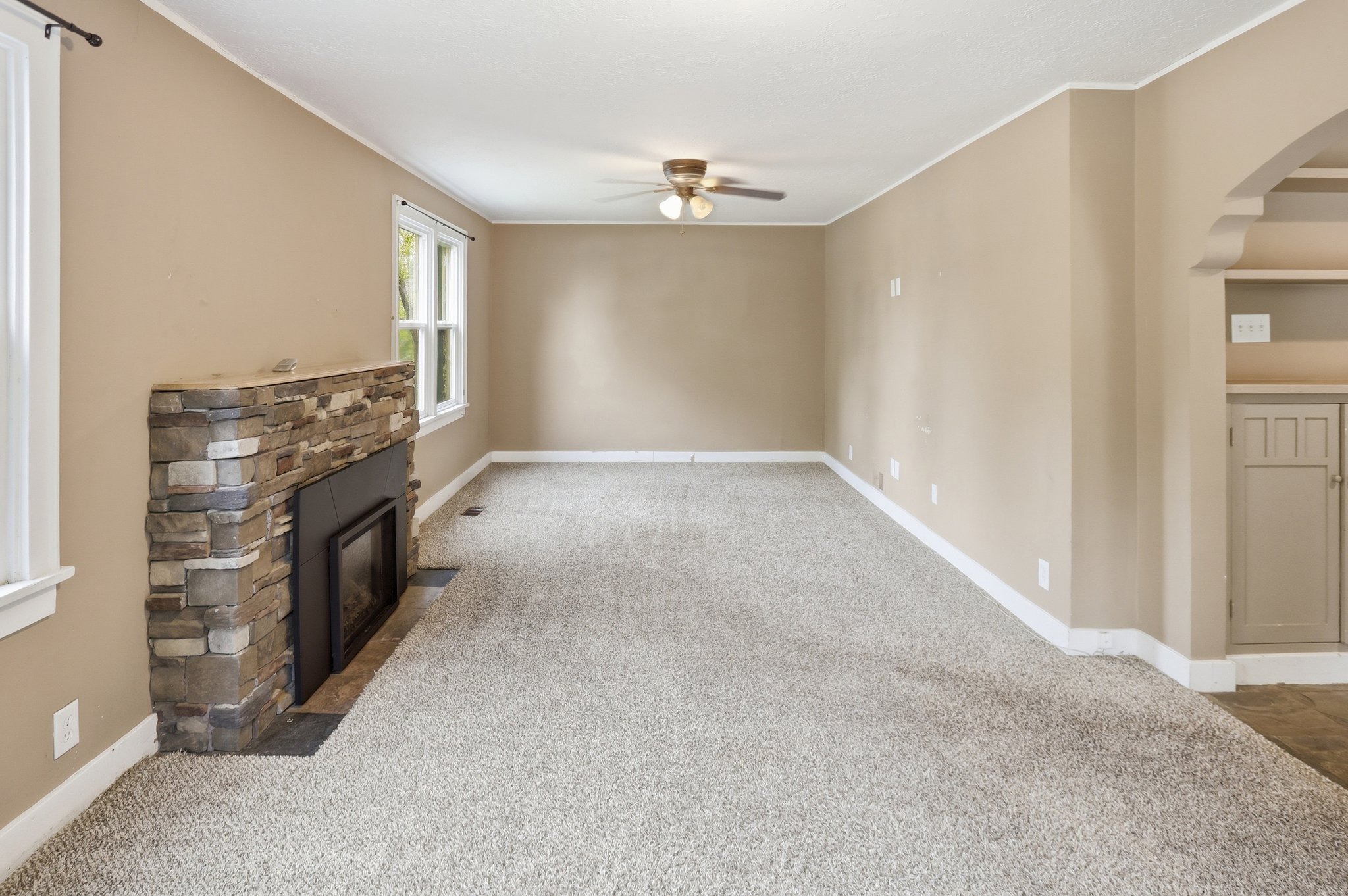 Unfurnished living room featuring a ceiling fan, carpet floors, crown molding, a stone fireplace, and arched walkways
