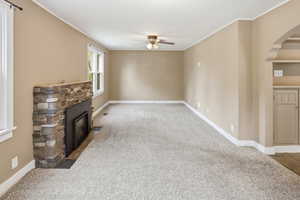 Unfurnished living room featuring a ceiling fan, carpet floors, crown molding, a stone fireplace, and arched walkways