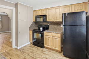 Kitchen with black appliances, light wood finish cabinetry, arched walkways, light wood-type flooring, and decorative backsplash
