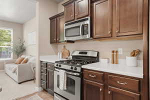 Kitchen with stainless steel appliances, light countertops, dark wood finish cabinetry, open floor plan, and light colored carpet