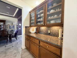 Bar area featuring wood finish cabinets, dark stone counters, glass fronted cabinets, a tray ceiling, and decorative backsplash