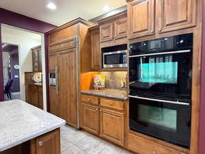Kitchen featuring built in appliances, decorative backsplash, wood finish cabinets, light tile patterned floors, and dark stone countertops