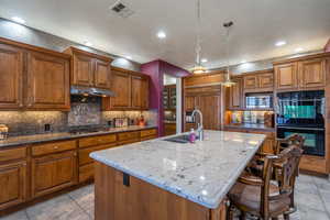 Kitchen with light stone countertops, stainless steel appliances, hanging light fixtures, wood finish cabinets, and an island with sink