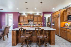 Kitchen with paneled refrigerator, backsplash, light stone countertops, decorative light fixtures, and a center island with sink