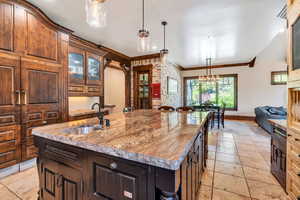 Kitchen featuring glass fronted cabinets, a center island with sink, ornamental molding, light stone counters, and stone tile flooring