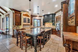 Two tone kitchen with glass insert cabinets, a kitchen breakfast bar, backsplash, a kitchen island with sink, and light stone counters