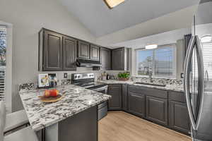 Kitchen with stainless steel appliances, light wood-style floors, vaulted ceiling, a peninsula, and light stone countertops