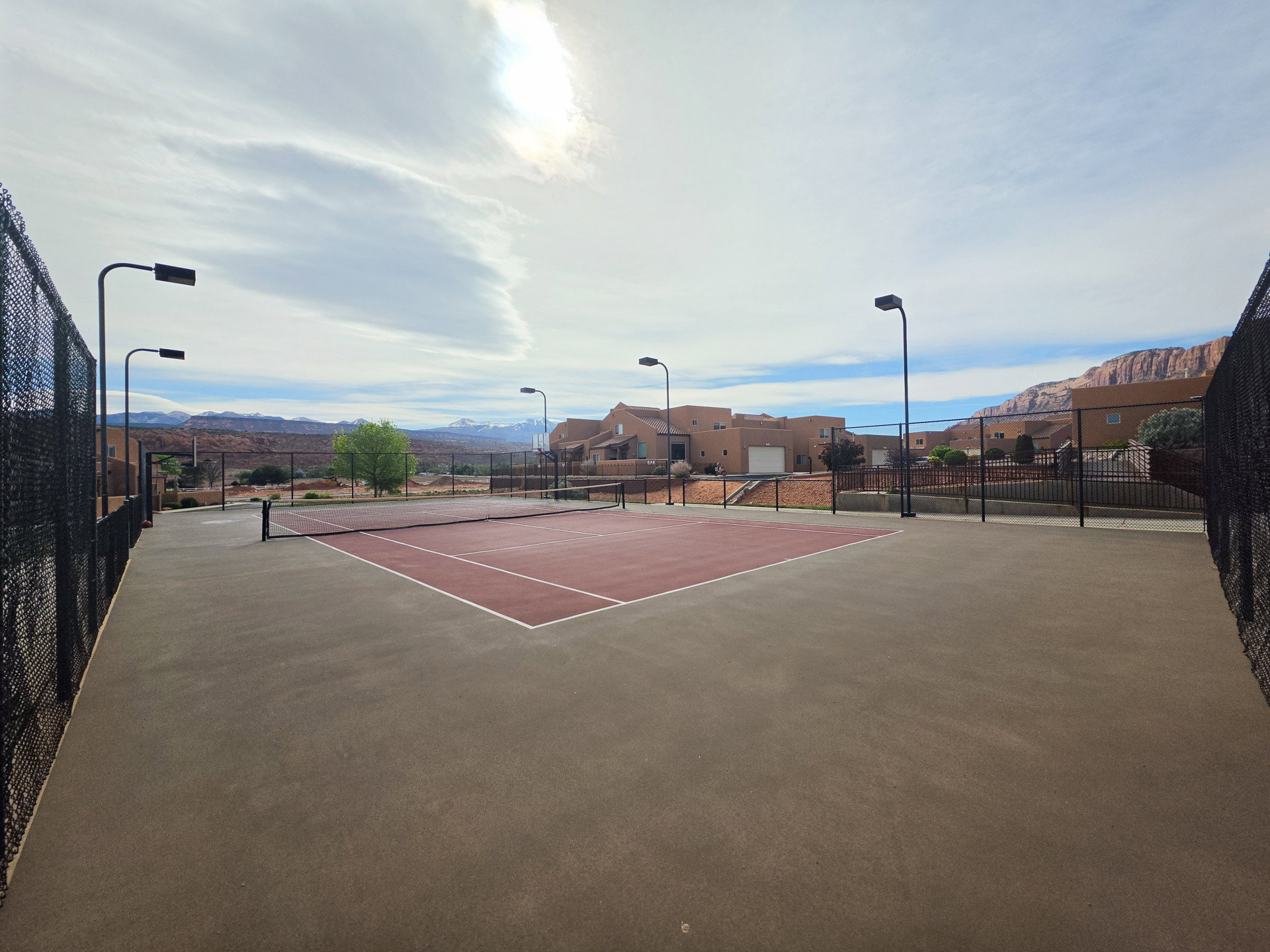 Outdoor tennis court with a red playing surface and white lines, surrounded by black chain-link fencing
