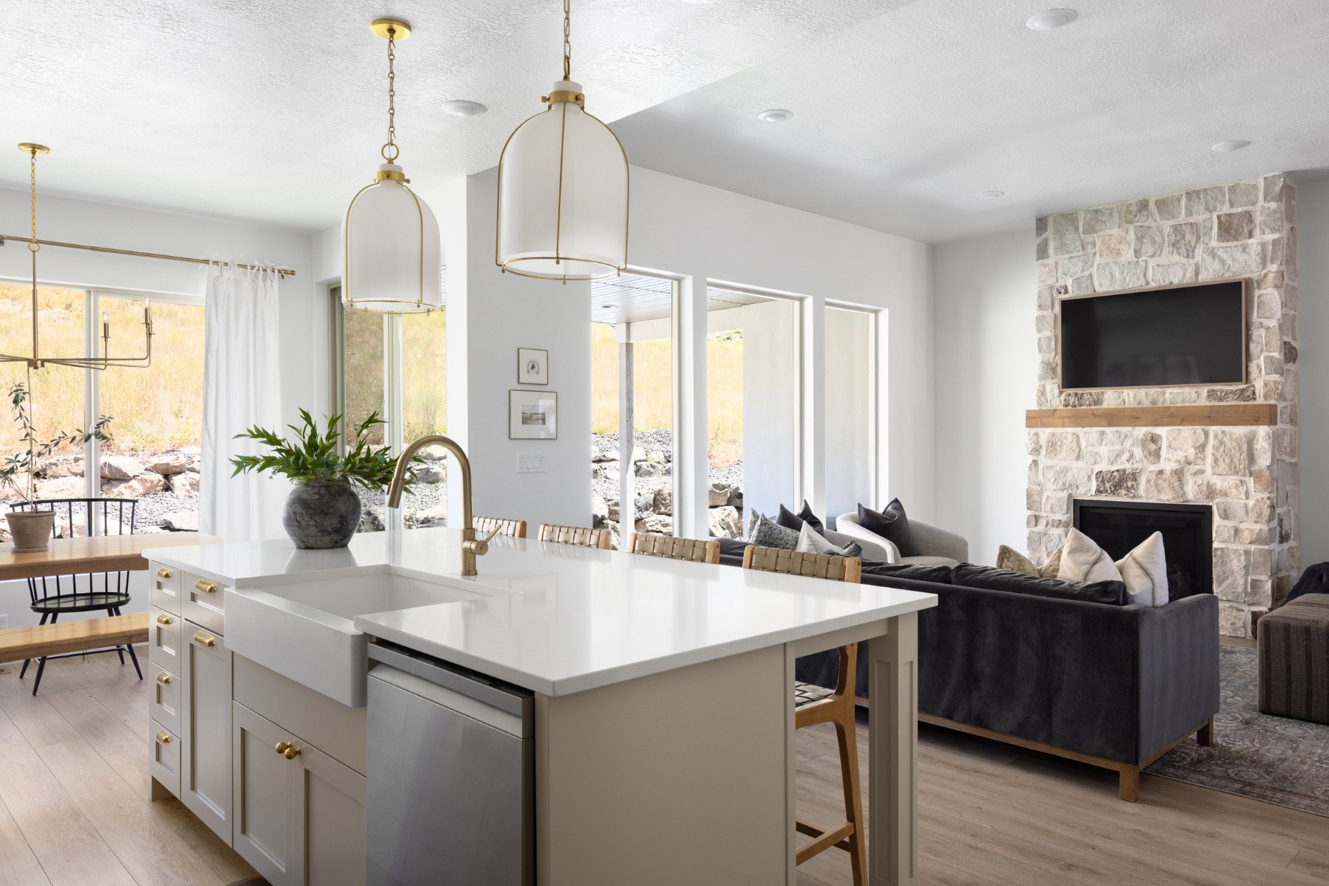 Open-concept living space featuring a stone-clad fireplace with wood mantel, large format windows, wood-finish flooring, and a kitchen island with a farmhouse sink and brass-finish fixtures