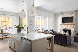Open-concept living space featuring a stone-clad fireplace with wood mantel, large format windows, wood-finish flooring, and a kitchen island with a farmhouse sink and brass-finish fixtures