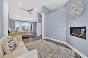Bedroom with vaulted ceiling, a glass covered fireplace, decorative columns, dark wood-style flooring, and a ceiling fan
