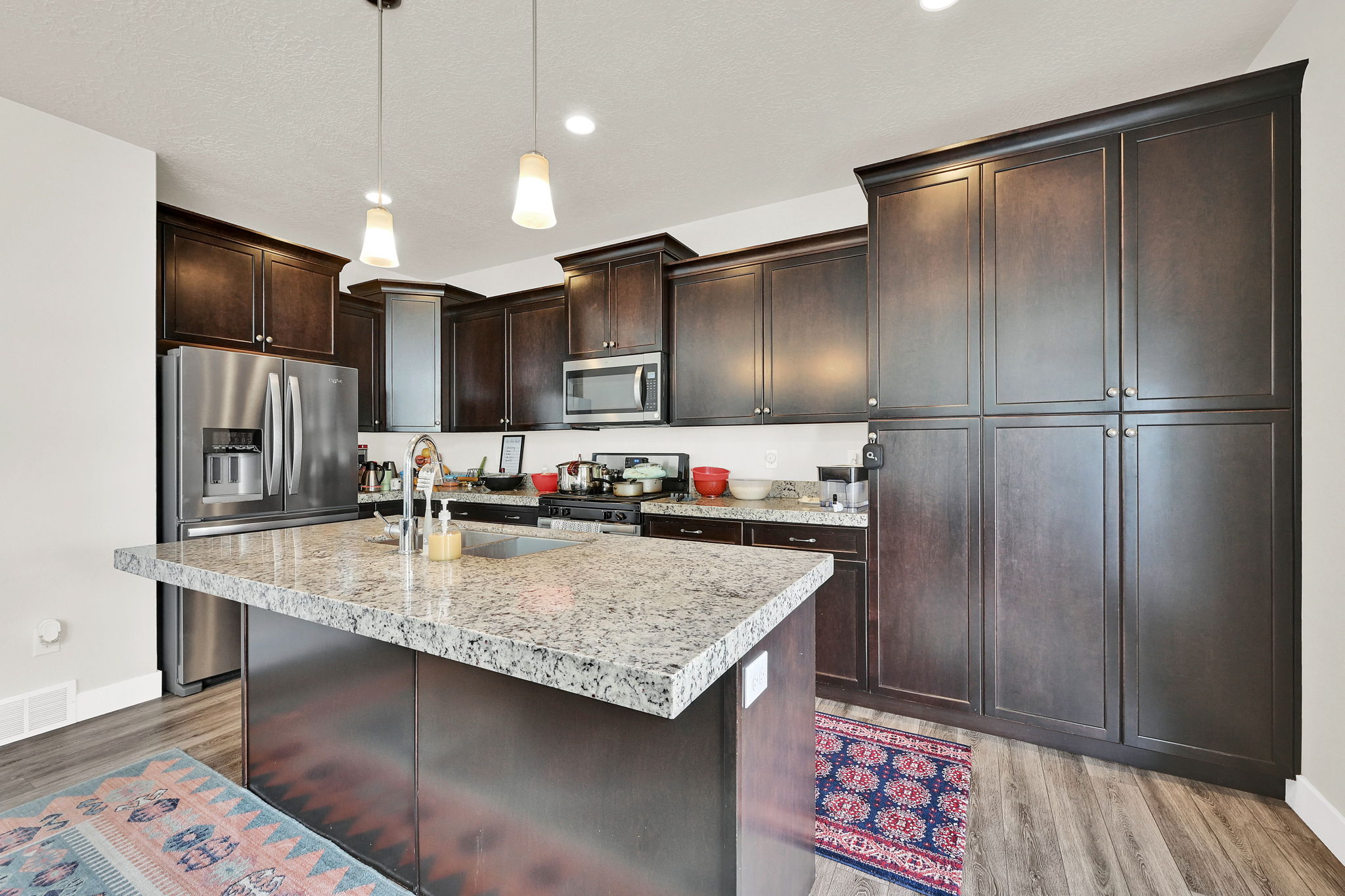 Kitchen featuring stainless steel appliances, a center island with sink, light wood finished floors, decorative light fixtures, and dark wood finish cabinets
