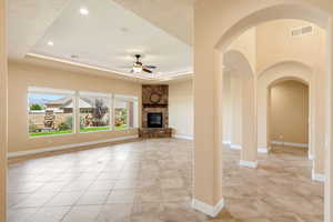 Unfurnished living room with a tray ceiling, a ceiling fan, a stone fireplace, arched walkways, and recessed lighting