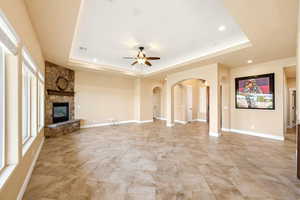 Unfurnished living room with arched walkways, a stone fireplace, ceiling fan, a raised ceiling, and recessed lighting