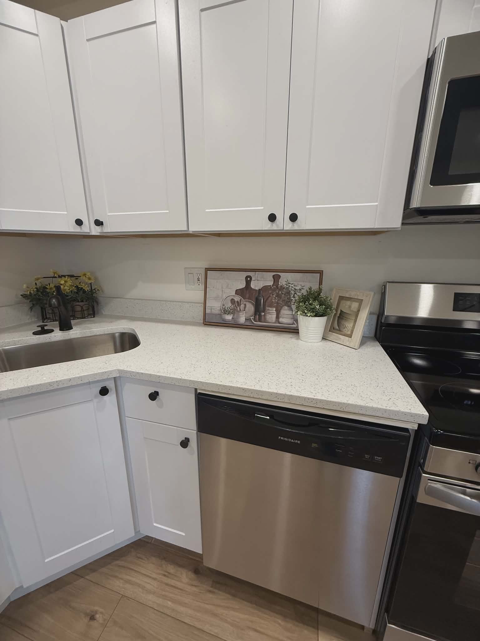Modern kitchen featuring white shaker cabinetry with dark hardware, speckled countertops, a stainless steel dishwasher, a stainless steel sink with a black faucet, and wood-finish flooring