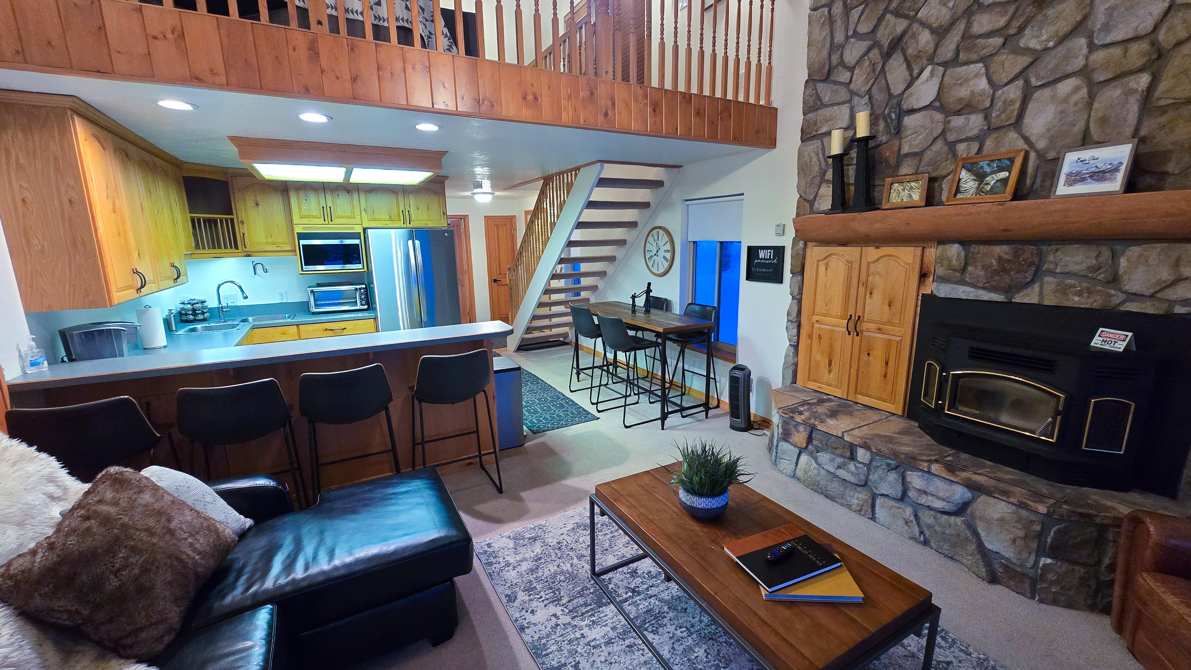 Open-concept living area featuring a stone-clad fireplace, wood-finish flooring, and a kitchen with wood cabinetry and stainless steel appliances