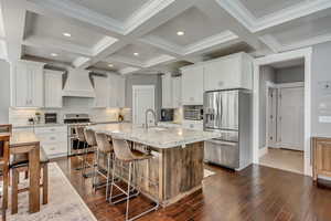 Kitchen featuring stainless steel appliances, an island with sink, dark wood finished floors, coffered ceiling, and light stone counters