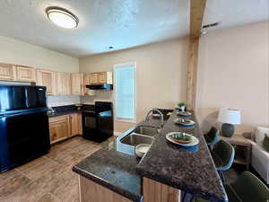 Kitchen featuring light wood finish cabinets, black appliances, dark stone countertops, and a textured ceiling