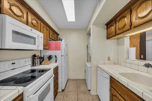 Kitchen with tile countertops, white appliances, wood finish cabinetry, light tile patterned floors, and washer / dryer