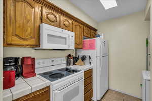 Kitchen with white appliances, tile countertops, wood finish cabinetry, light tile patterned floors, and washer / clothes dryer