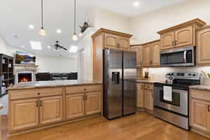 Kitchen featuring stainless steel appliances, pendant lighting, a warm lit fireplace, light stone countertops, and light wood-style flooring