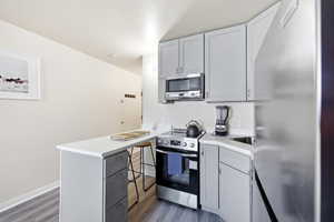 Kitchen featuring stainless steel appliances, a peninsula, gray cabinetry, a kitchen bar, and dark wood-style flooring
