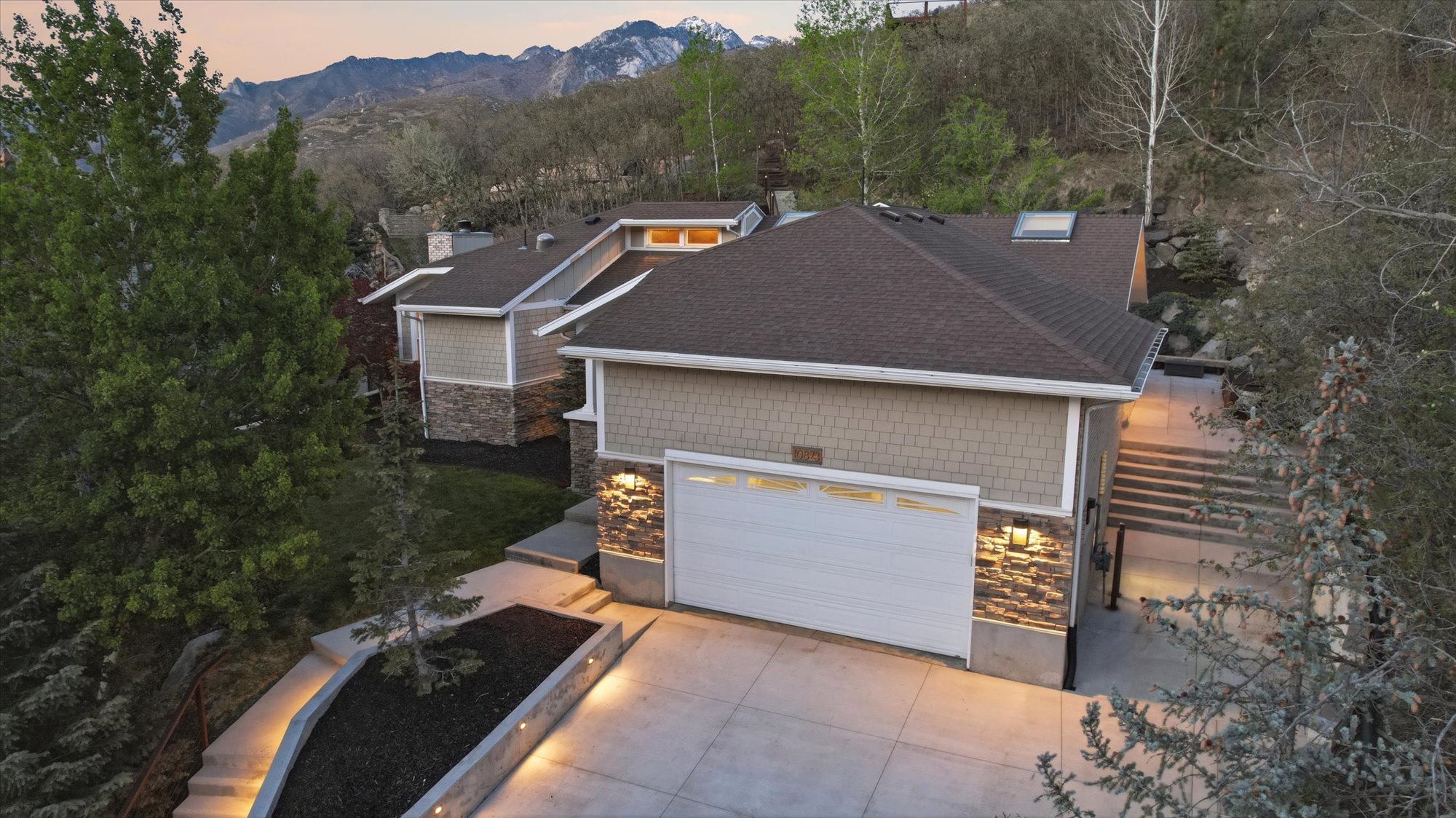 View of front of property featuring concrete driveway, stone siding, roof with shingles, a mountain view, and view of wooded area