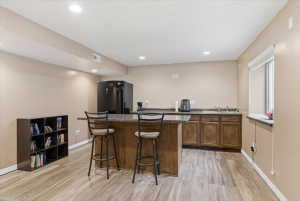 Indoor wet bar with freestanding refrigerator, light wood-type flooring, wood finish cabinetry, dark stone countertops, and recessed lighting