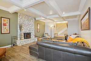 Living room featuring a stone fireplace, wood-type flooring, crown molding, and coffered ceiling