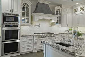 Kitchen featuring glass insert cabinets, stainless steel appliances, white cabinetry, light stone countertops, and backsplash