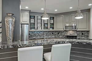 Kitchen with stainless steel appliances, tasteful backsplash, glass insert cabinets, hanging light fixtures, and dark stone countertops