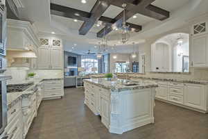 Kitchen featuring backsplash, glass insert cabinets, coffered ceiling, a center island with sink, and light stone counters