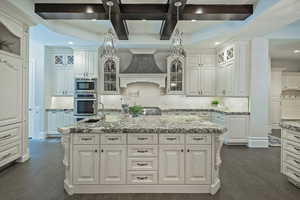 Kitchen featuring glass fronted cabinets, coffered ceiling, dark wood-style flooring, light stone counters, and a center island with sink