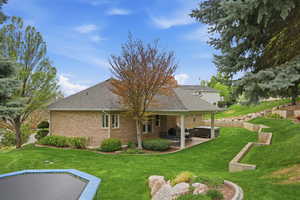 Back of house with a patio area, brick siding, a hot tub, roof with shingles, and a lawn