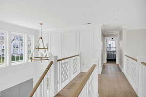 Hall featuring a decorative wall, suspended lighting, light wood-style floors, ornamental molding, and an upstairs landing
