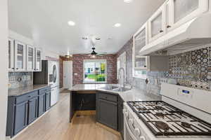 Two tone kitchen with glass insert cabinets, white range with gas stovetop, a peninsula, light wood-style flooring, and light stone counters