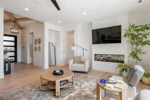 Living area with light wood finished floors, beam ceiling, a glass covered fireplace, suspended lighting, and ceiling fan