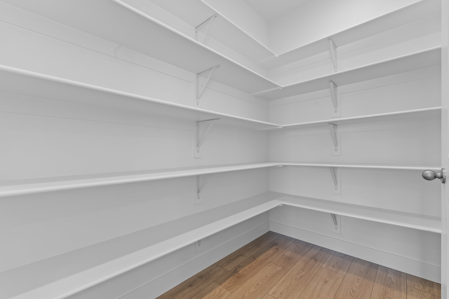 Walk-in pantry featuring abundant white shelving, wood-finish flooring, and a white door with a brushed nickel doorknob