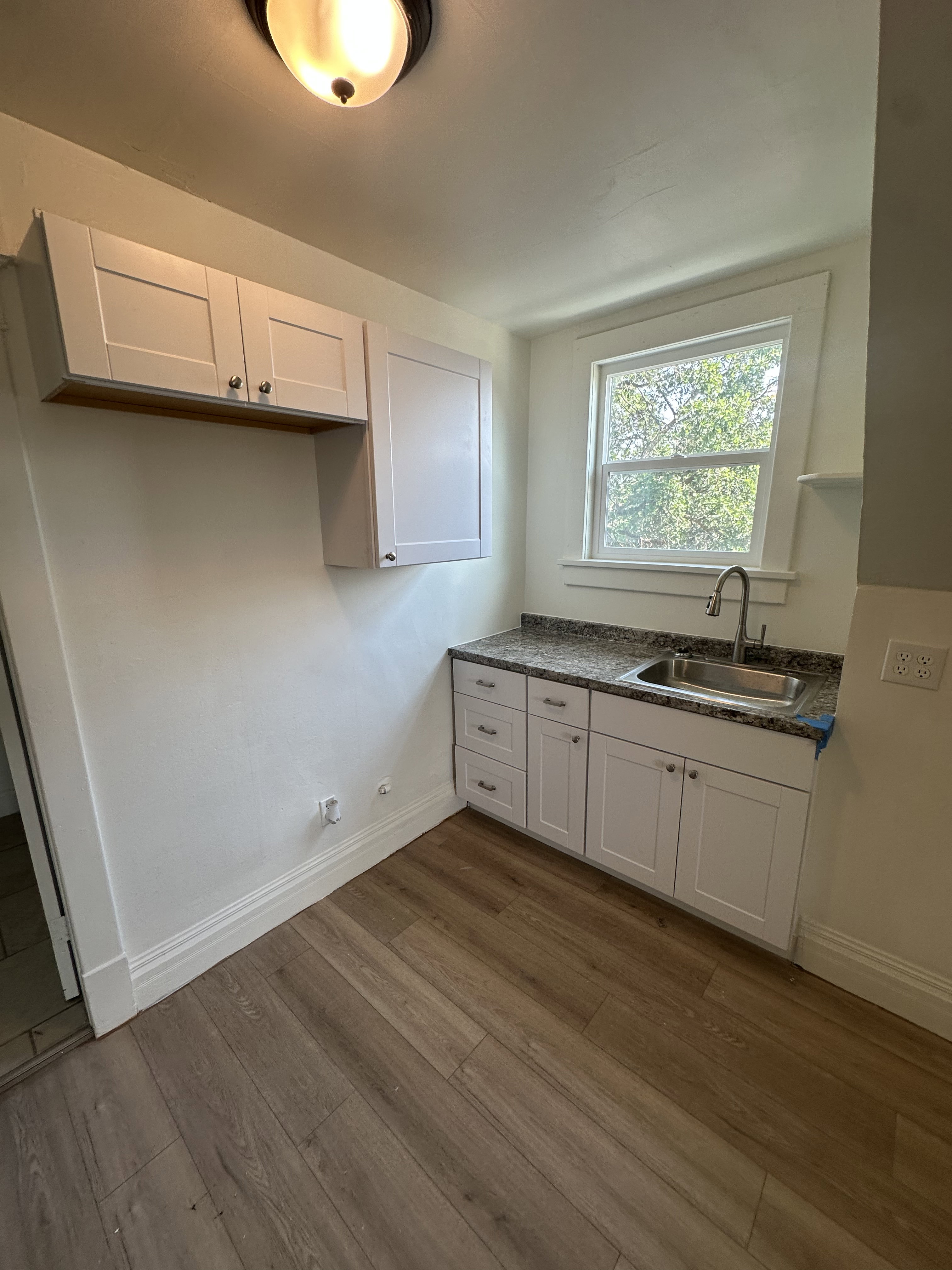Compact kitchen area featuring wood-finish flooring, white shaker-style cabinetry, a stainless steel sink, and speckled countertops