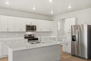 Kitchen featuring stainless steel appliances, a kitchen island with sink, white cabinets, light stone counters, and light wood-style flooring
