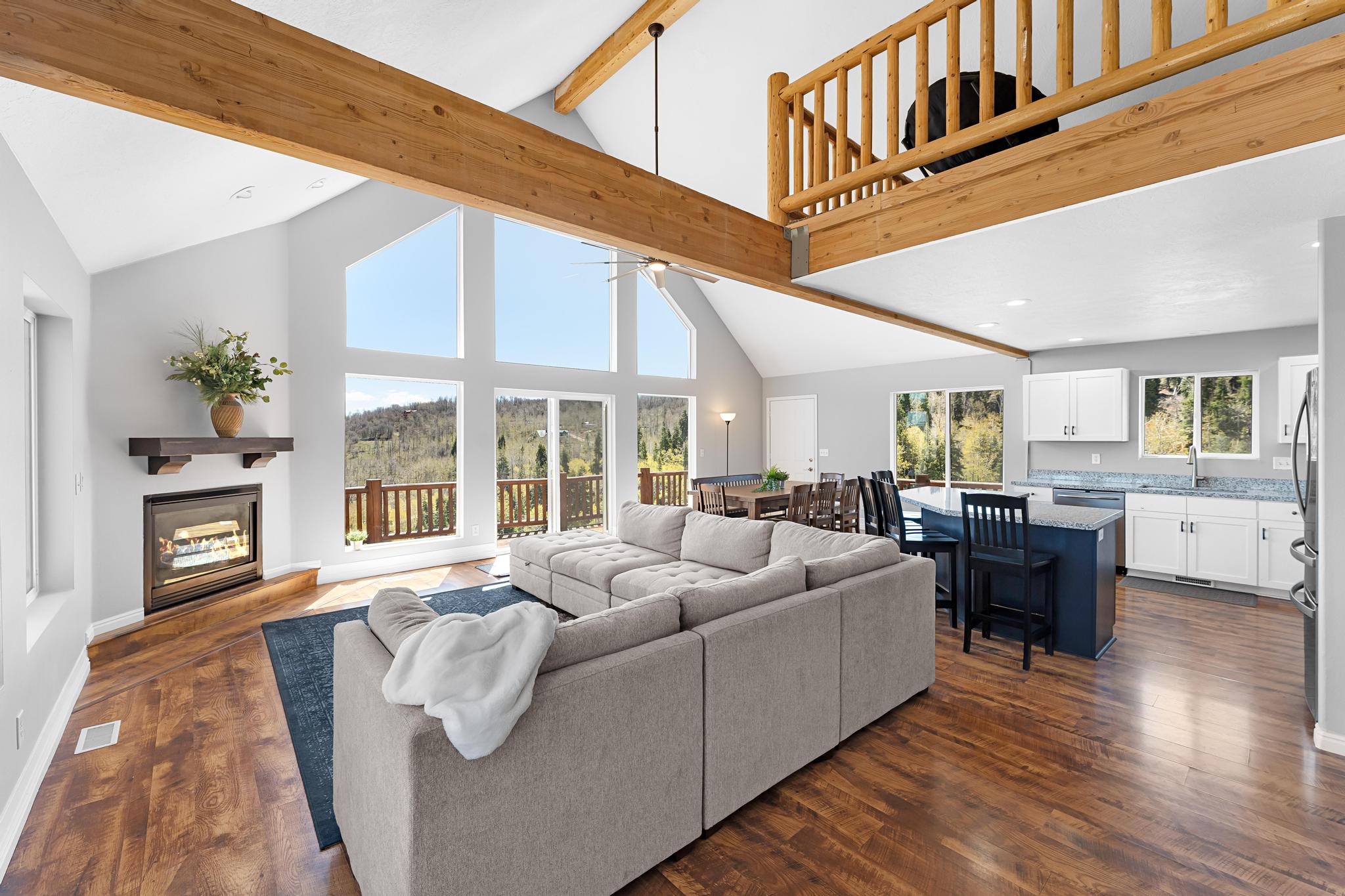 Open-concept living space featuring vaulted ceilings with exposed wood beams, a stone fireplace with a wood mantle, and large windows