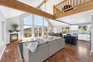 Open-concept living space featuring vaulted ceilings with exposed wood beams, a stone fireplace with a wood mantle, and large windows