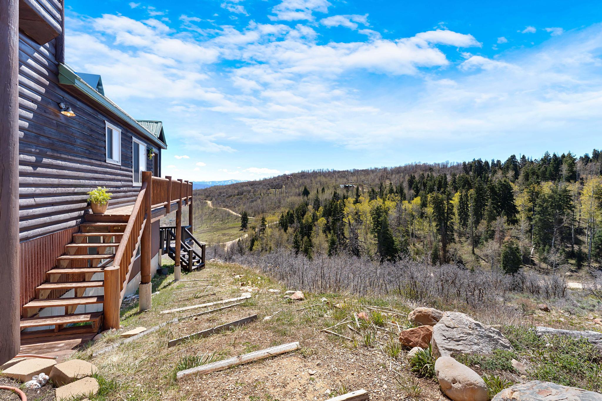 Log cabin exterior featuring a multi-level deck, wood stairs, and natural landscaping with mature trees and distant hills