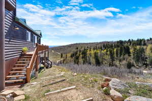 Log cabin exterior featuring a multi-level deck, wood stairs, and natural landscaping with mature trees and distant hills