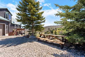 Gravel driveway with a multi-level deck, surrounding mature evergreen trees, and a mountain backdrop